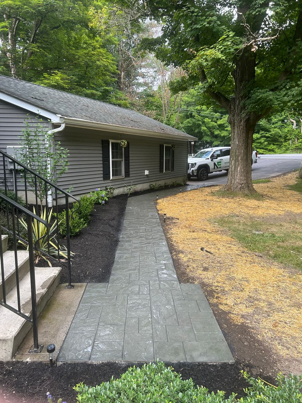 Pathway leading to a house, bordered by landscaping and a parked vehicle in a wooded area.
